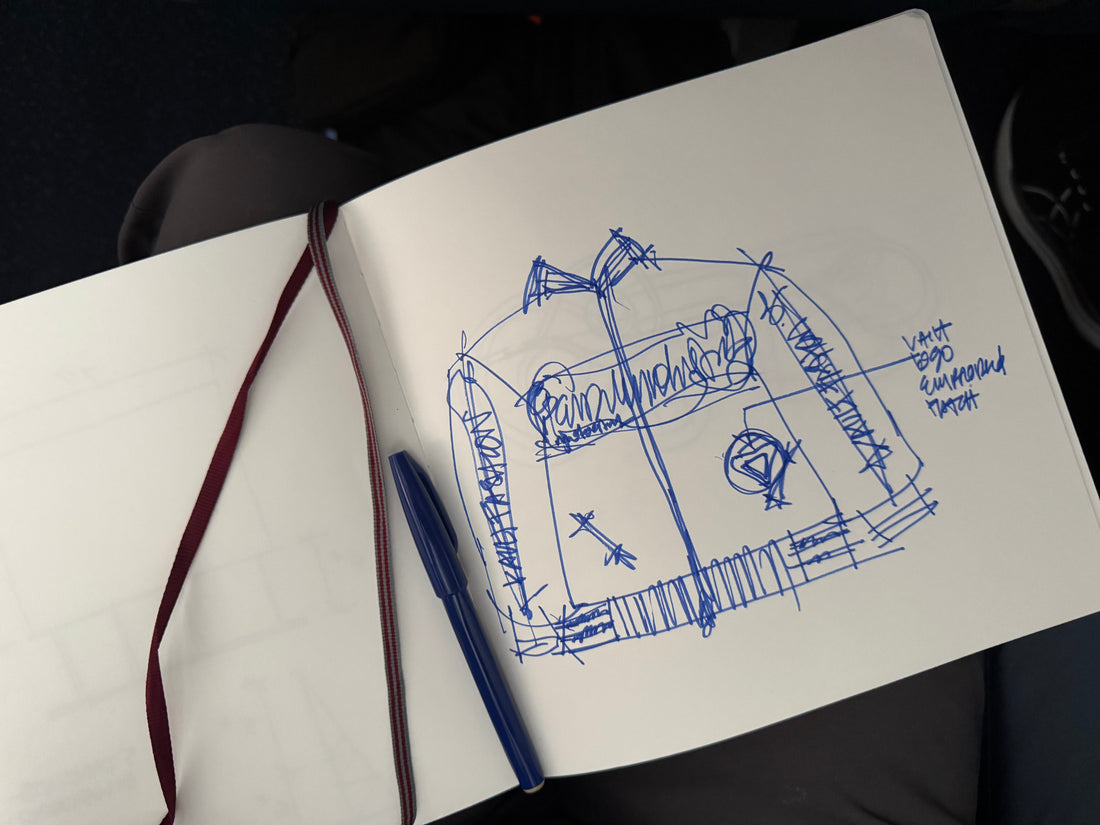 Hand-drawn blue ink sketch of a hoodie design in an open notebook with notes and placement for “Vault” logo and embroidered patch, captured alongside a blue pen and burgundy bookmark ribbon.