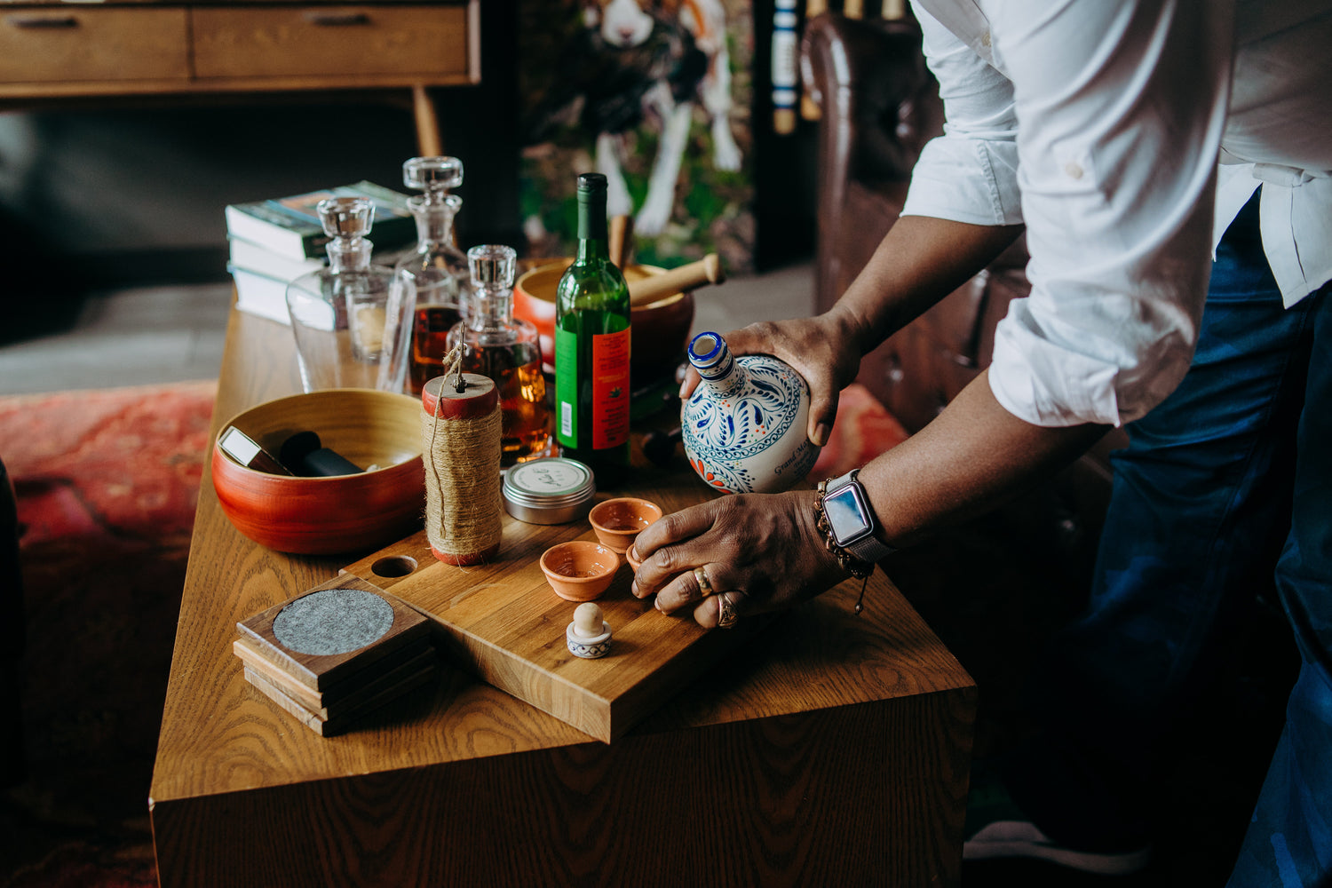 Close-up of a man pouring tequila into ceramic cups on a wooden table set with bottles, decanters, and artisanal barware. A cozy, curated interior with rich textures, handcrafted objects, and a warm, refined atmosphere.