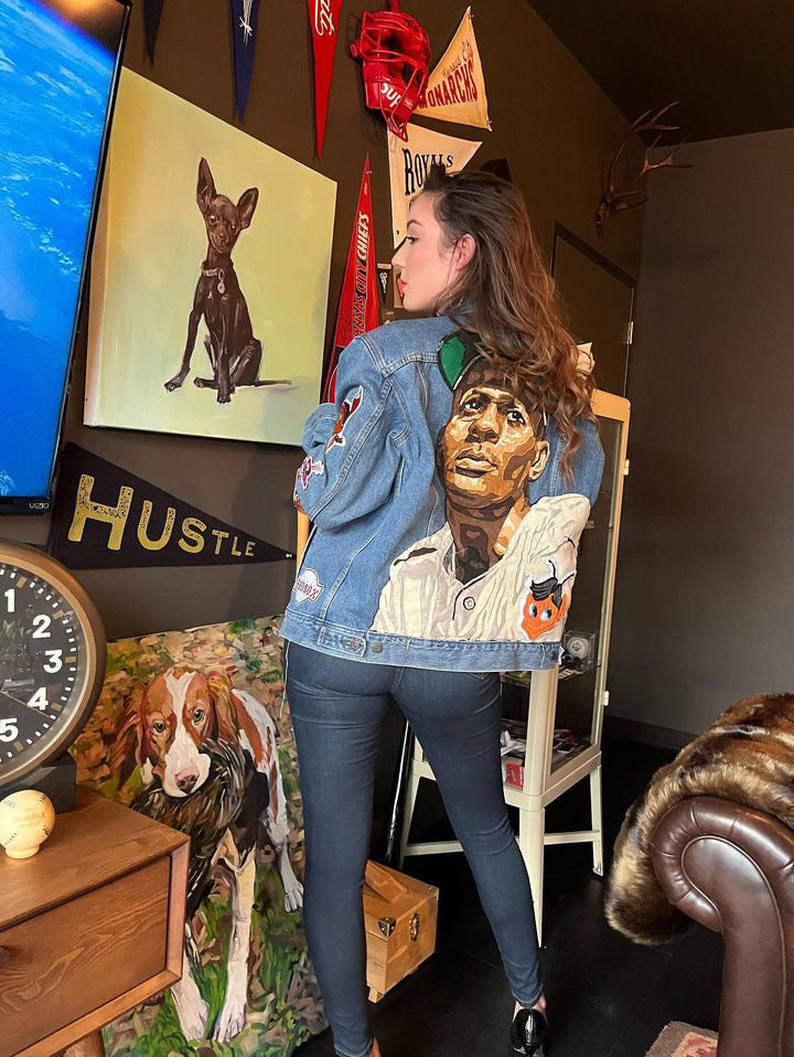 Woman poses in a custom denim jacket featuring a painted portrait of a baseball player on the back. Surrounded by sports memorabilia and dog art, the setting blends athletic heritage with creative expression and personalized streetwear flair.