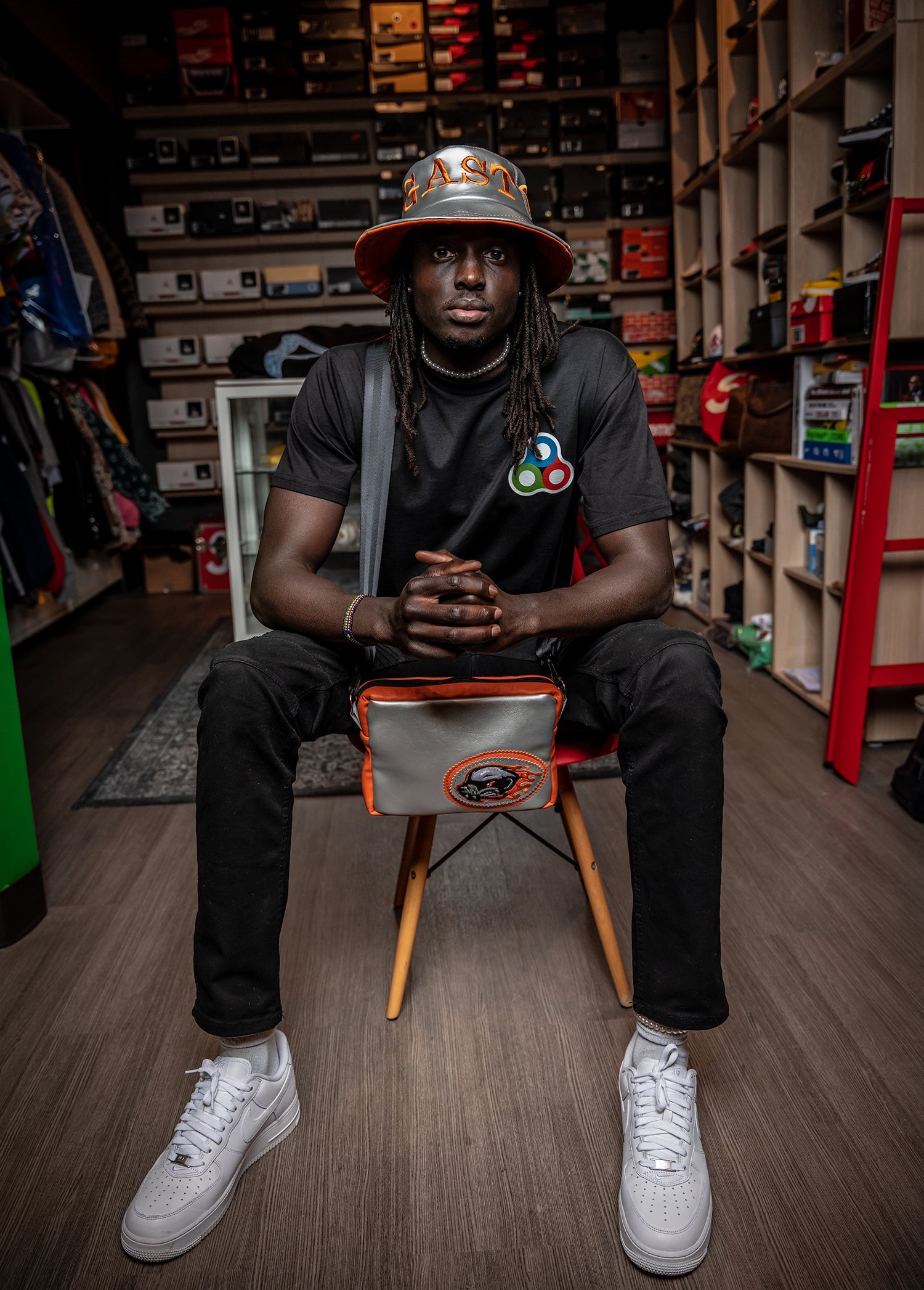 Stylish man seated in a sneaker boutique wearing a black tee with a colorful graphic, black jeans, white sneakers, and a leather bucket hat labeled “GAST.” Orange and gray crossbody bag with embroidered patch adds a bold accent to the look.