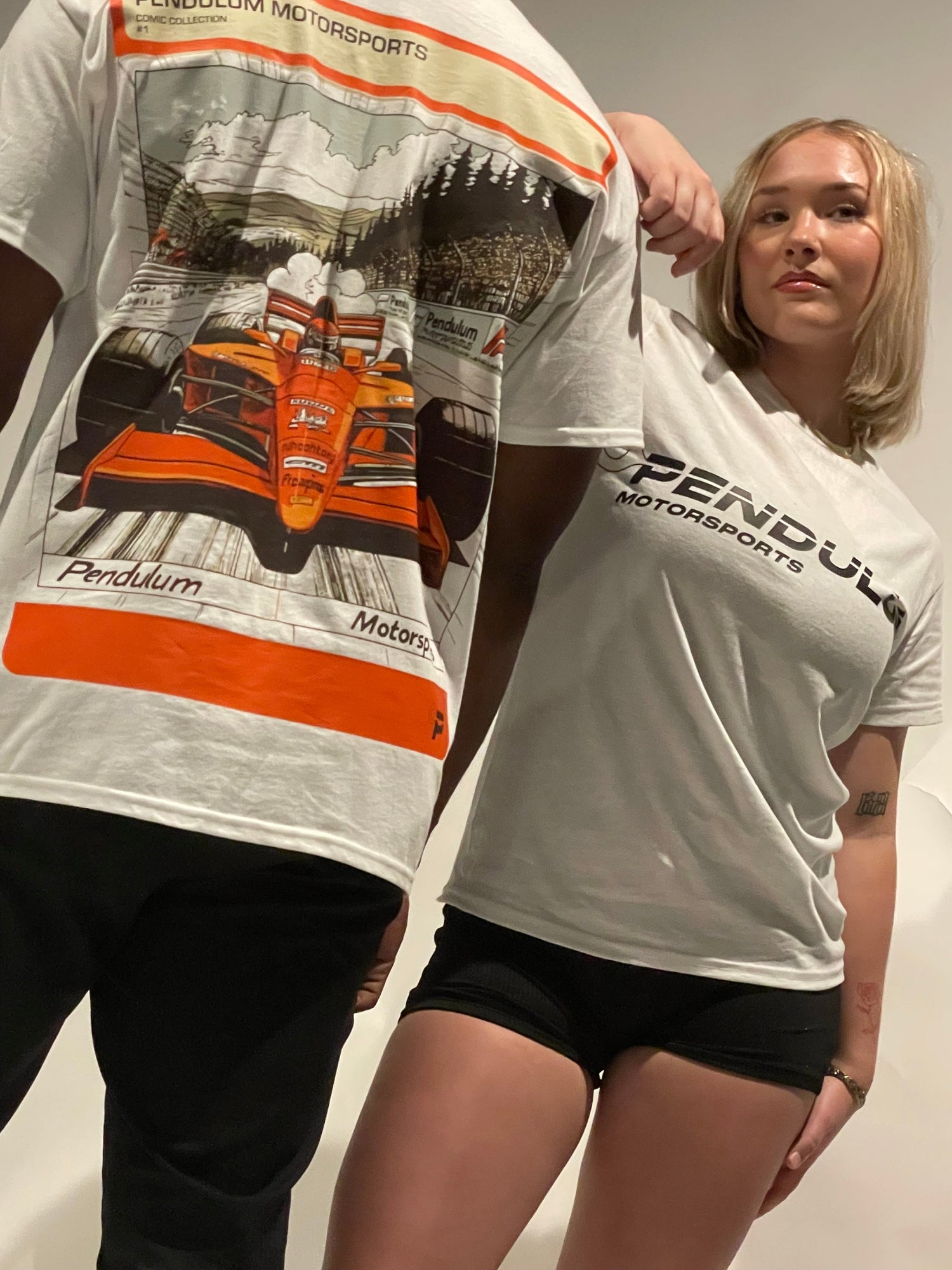 Two models wear Pendulum Motorsports tees—one shows the back with a vintage race car graphic in red and orange, the other wears the front-logo version. Streetwear style blending motorsport heritage with modern graphic design.
