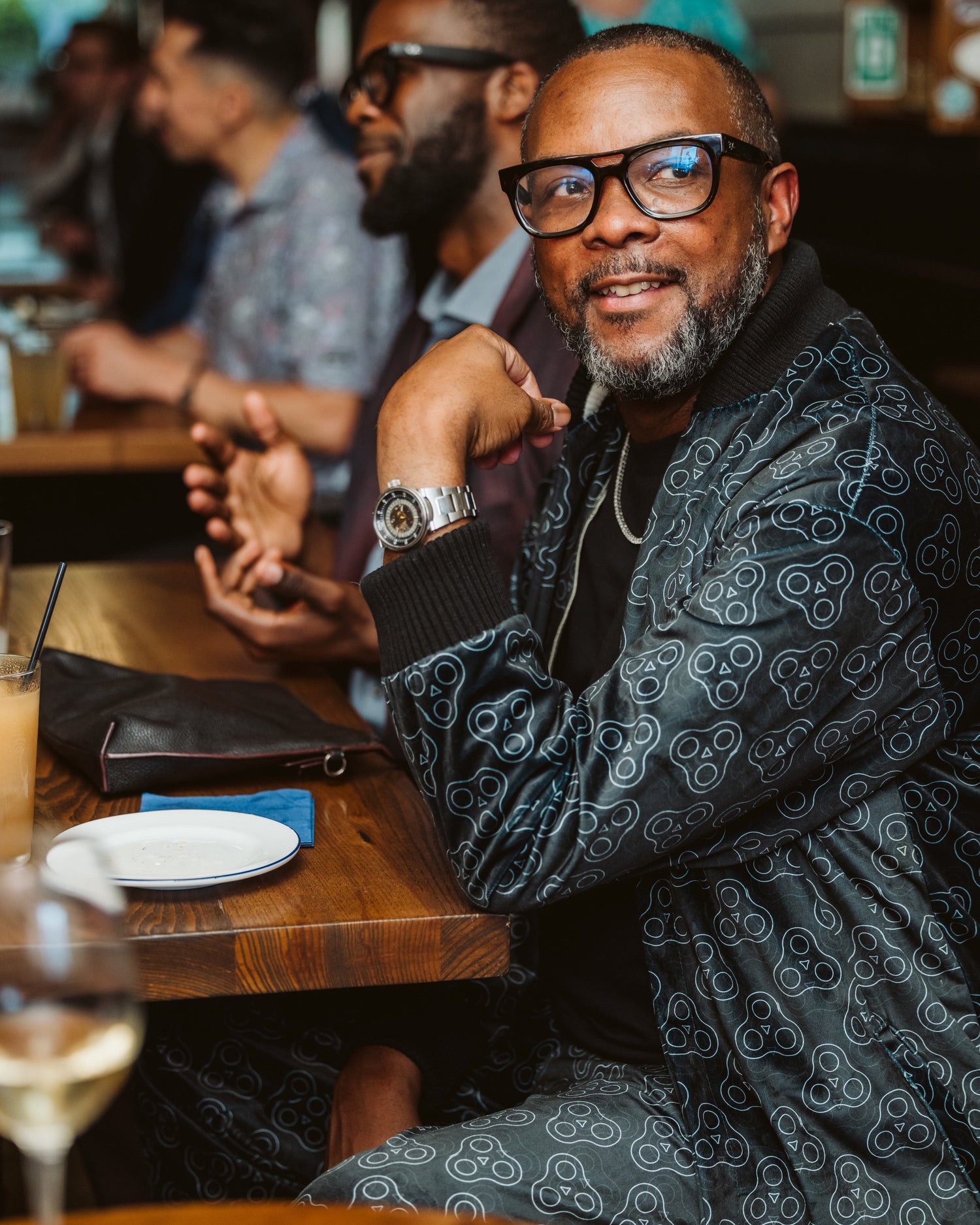 Man seated at a restaurant wearing a matching black patterned jacket and pants set with minimalist skull graphics. Stylish look accented by oversized glasses, silver watch, and a confident expression. Casual luxury with bold streetwear flair.