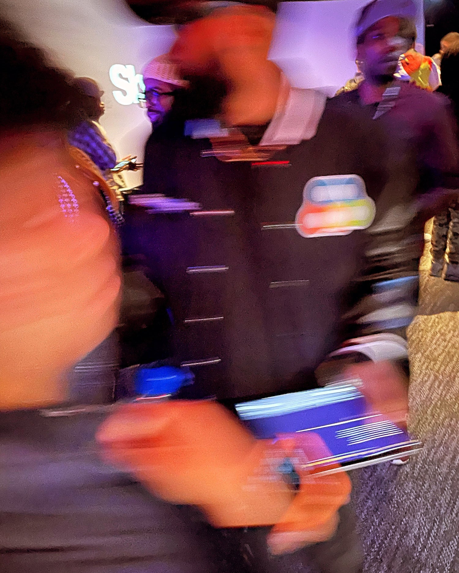 Blurred motion photo of a crowded event with people in streetwear and bold patches, capturing energy and movement. A man in focus wears a black jacket with a colorful embroidered patch, reflecting a dynamic urban fashion scene.