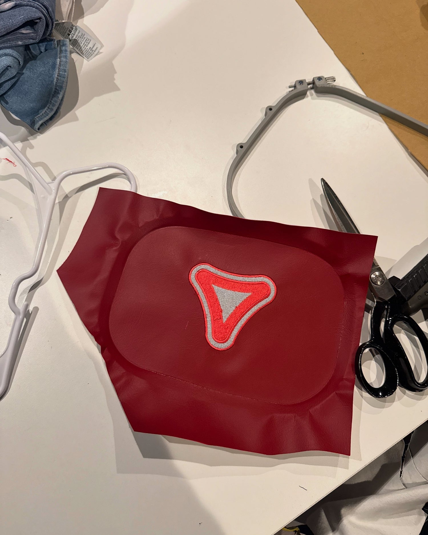 Close-up of a red leather panel with an embroidered triangular patch in red, white, and gray. Surrounded by scissors, hanger, and denim, the image captures a behind-the-scenes look at the design and crafting process of a custom fashion piece.