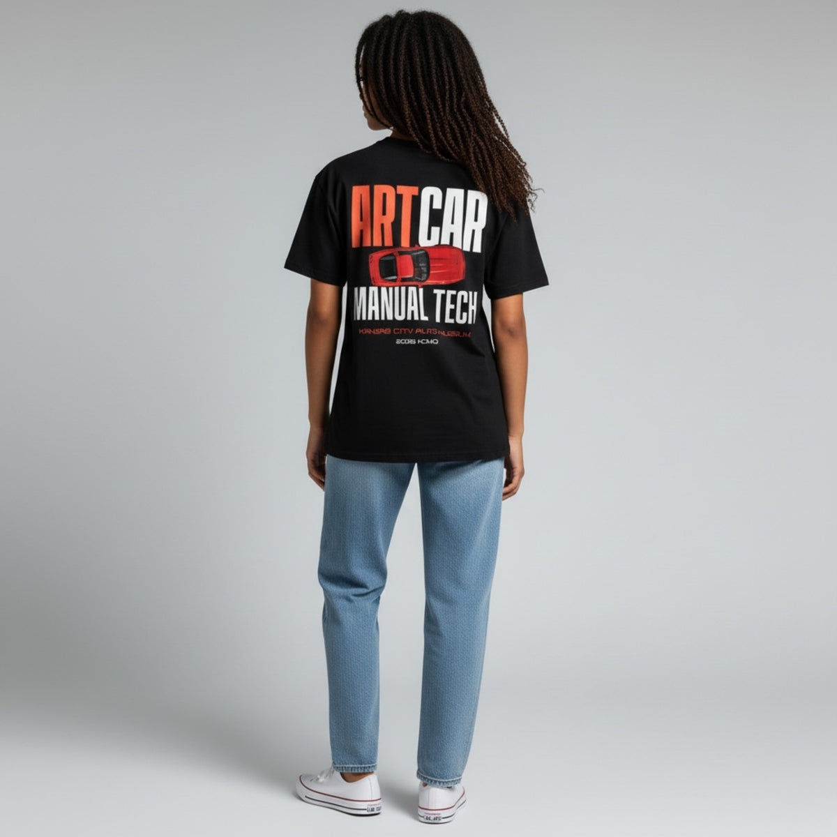 A model wearing the black KCAM Art Car tee seen from behind, showing the full ARTCAR MANUAL TECH back print with a red art car above Kansas City Auto Museum, 2026 KCMO