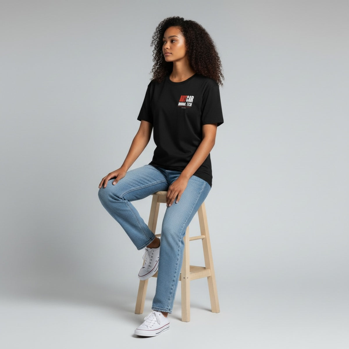 A model seated on a stool wearing the black KCAM Art Car tee, front view, with the ARTCAR MANUAL TECH print on the left chest