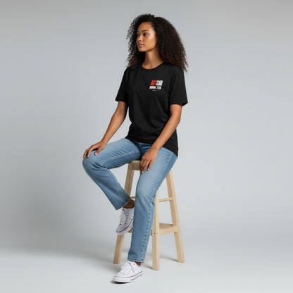 A model seated on a stool wearing the black KCAM Art Car tee, front view, with the ARTCAR MANUAL TECH print on the left chest